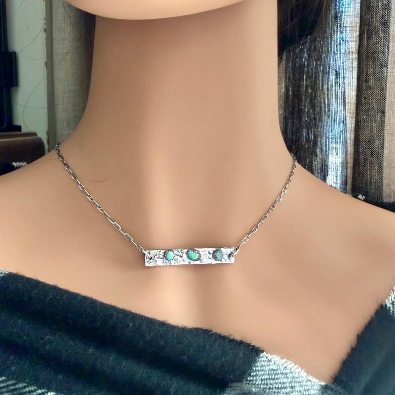 May include: A silver necklace featuring a rectangular pendant with three turquoise-colored stones. The pendant is attached to a delicate chain. The necklace is worn on a person's neck, against a black and white plaid shirt.