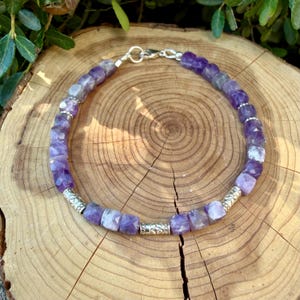 Puede incluir: Una pulsera de cuentas compuesta por cubos de amatista morada facetada dispuestos en un patr&oacute;n circular, intercalados con peque&ntilde;as cuentas espaciadoras cil&iacute;ndricas de metal plateado y un cierre de mosquet&oacute;n de metal plateado.