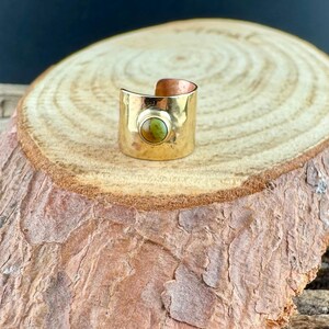 May include: A wide, gold-colored ring with a hammered texture and a copper interior. The ring features a round, olive-green stone set in the center. The ring is displayed on a wooden surface.