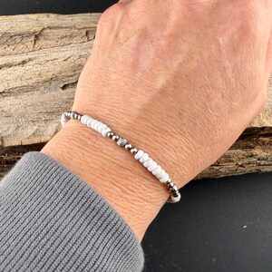 May include: A white beaded bracelet with silver-colored accents. The bracelet features small, round white beads interspersed with textured silver beads. The bracelet is worn on a wrist, with a gray sleeve in the foreground and a wooden background.
