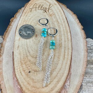 May include: A pair of silver hoop earrings with turquoise beads and dangling silver chains. The earrings are displayed on a wooden surface with the text "Ally K" and a buffalo nickel coin.