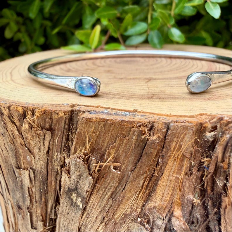 K&ouml;nnte beinhalten: Ein silberfarbenes Armreif-Armband mit ovalen, irisierenden Edelsteinen an jedem Ende. Das Armband wird auf einer Holzoberfl&auml;che mit gr&uuml;nem Laub im Hintergrund pr&auml;sentiert. Die Edelsteine haben eine blaue und wei&szlig;e Farbe.