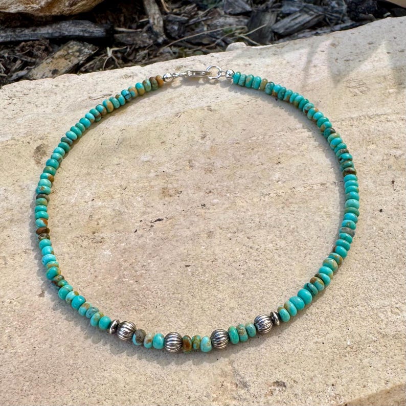 May include: A turquoise beaded necklace with silver accents. The necklace features small, round turquoise beads in shades of blue and green, interspersed with silver beads. The necklace is secured with a silver clasp.