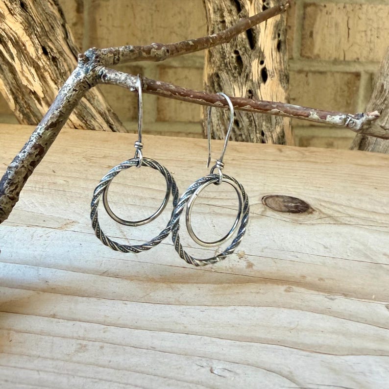 May include: A pair of silver-toned dangle earrings featuring a double-hoop design. Each earring consists of a larger, twisted-texture outer hoop and a smaller, smooth inner hoop, suspended from a simple curved ear wire hook for a secure fit.