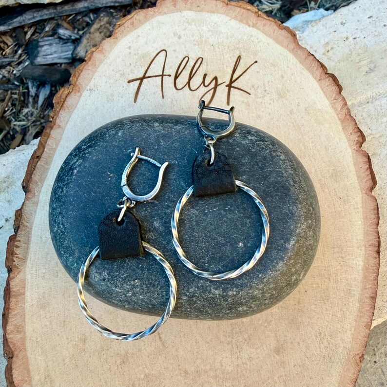 May include: A pair of silver hoop earrings with a twisted design. Each earring features a small black leather accent and a hinged clasp. The earrings are displayed on a dark gray stone, with the name "Allyk" written in cursive on a wooden surface.