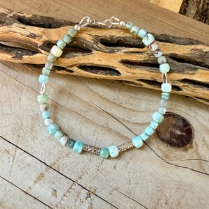Puede incluir: Una pulsera de cuentas con peque&ntilde;as piedras preciosas facetadas en tonos azul claro y verde, intercaladas con dos espaciadores met&aacute;licos cil&iacute;ndricos de color plateado. La pulsera se asegura con un cierre de mosquet&oacute;n y una anilla plateada.