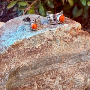 May include: Two silver-toned metal rings featuring a wide, grooved band design. Each ring is set with a single, round, vibrant orange stone at the center. The metal has a polished finish with horizontal lines etched into the surface of the bands.