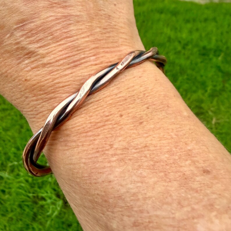 May include: A copper-colored twisted metal bracelet. The bracelet is made of two strands of metal that are intertwined. The bracelet is worn on a wrist, with a green background.