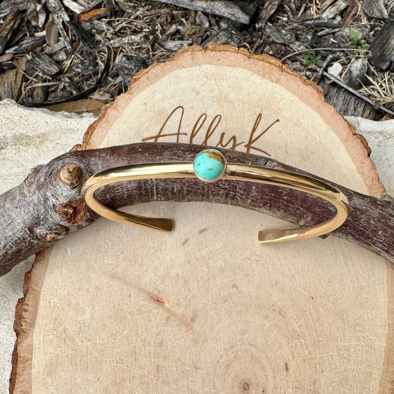 Op de afbeelding: Een goudkleurige manchetarmband met een turkooizen steen in het midden. De armband wordt gepresenteerd op een houten oppervlak met het handgeschreven woord "Allyk". De armband heeft een eenvoudig, open ontwerp.