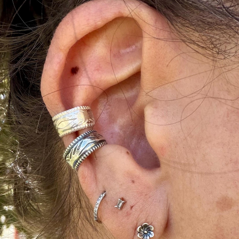 May include: Two silver-toned ear cuffs with etched patterns stacked on the upper cartilage, accompanied by a small silver hoop earring, a tiny rectangular stud, and a small silver flower-shaped stud on the earlobe.