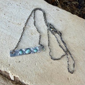 May include: A silver-toned metal necklace featuring a rectangular bar pendant with an intricate etched swirl pattern. The bar is set with three smooth, oval-shaped light blue-green stones. The pendant hangs from a delicate, thin metal chain.