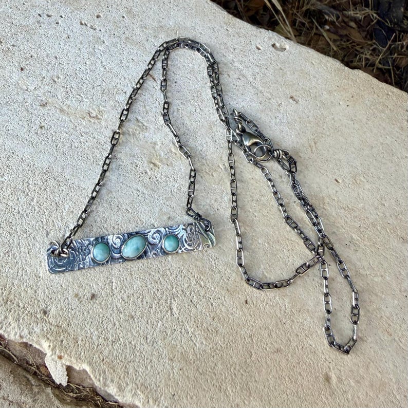 May include: A silver-toned metal necklace featuring a rectangular bar pendant with an intricate etched swirl pattern. The bar is set with three smooth, oval-shaped light blue-green stones. The pendant hangs from a delicate, thin metal chain.
