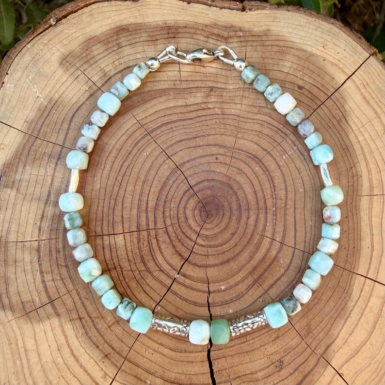 Puede incluir: Una pulsera de cuentas con peque&ntilde;as piedras cuadradas de color verde claro y blanco, intercaladas con dos espaciadores cil&iacute;ndricos de metal plateado texturizado. La pulsera se asegura con un cierre de mosquet&oacute;n de metal plateado.