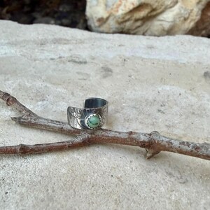 May include: A silver-toned ring with an open design, featuring a light green gemstone. The ring has a textured band with floral detailing. The ring is resting on a small, brown branch.