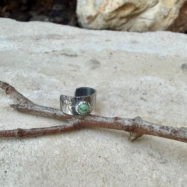 May include: A silver-toned ring with an open design, featuring a light green gemstone. The ring has a textured band with floral detailing. The ring is resting on a small, brown branch.
