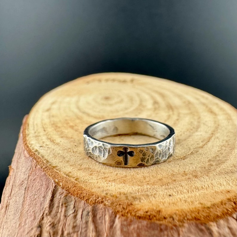 May include: A silver ring with a hammered texture and a dark cross design. The ring is set on a wooden surface, showcasing its handcrafted details. The band has a textured surface and a dark cross in the center.