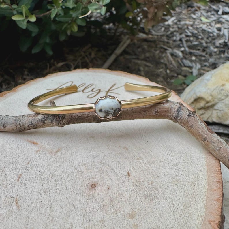 May include: A gold-colored cuff bracelet with a central, round, white and gray stone. The bracelet is resting on a small branch, with a wooden surface in the background. The bracelet has a simple, elegant design.