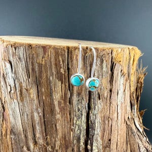 May include: A pair of drop earrings featuring round, polished turquoise stones with brown matrix veining. Each stone is set in a smooth silver-toned metal bezel and attached to a curved silver-toned ear wire for a simple, dangling design.