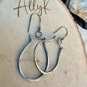 May include: A pair of silver-toned teardrop earrings with textured detailing. The earrings have a hook closure and are displayed on a wooden surface. The word "Allyk" is handwritten in brown ink in the upper left corner.