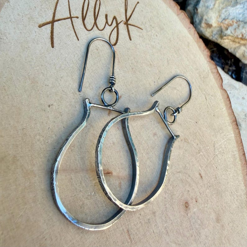 May include: A pair of silver-toned teardrop earrings with textured detailing. The earrings have a hook closure and are displayed on a wooden surface. The word "Allyk" is handwritten in brown ink in the upper left corner.