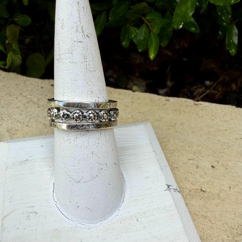 May include: A silver ring with a floral design is displayed on a white ring holder. The ring has three bands, with the middle band featuring a repeating pattern of small flowers. The ring is made of a shiny, reflective metal.