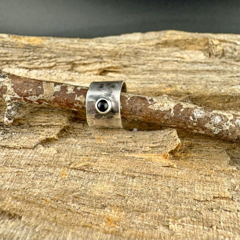 May include: A silver-toned ring with a hammered texture and a black gemstone. The ring is set against a natural wood background, showcasing its handcrafted design. The ring's band is approximately 0.75 inches wide.