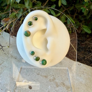 May include: A set of six round stud earrings featuring green and brown mottled stones set in silver-toned metal. The earrings are displayed on a white ear-shaped model, with three studs along the upper cartilage and three along the earlobe.