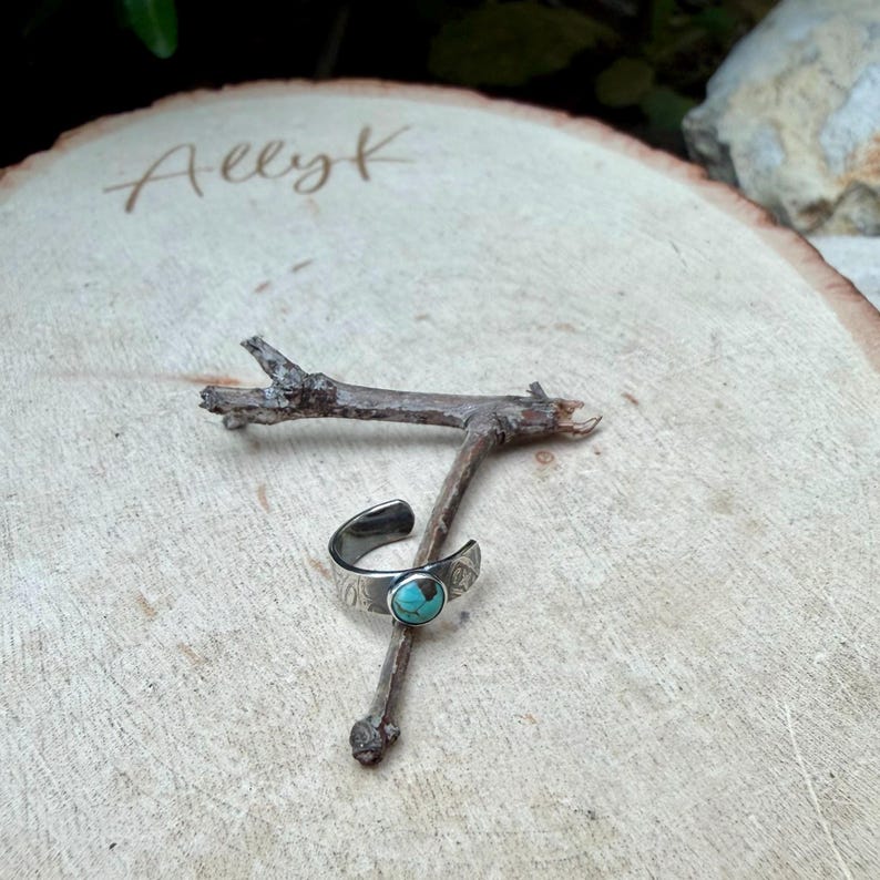 K&ouml;nnte beinhalten: Ein silberfarbener, verstellbarer Ring mit einem t&uuml;rkisfarbenen Stein, der auf einem kleinen Zweig liegt. Der Ring hat ein dekoratives Muster und ist vor einem hellen Holzgrund platziert. Das Wort "Allyk" steht auf dem Holz.