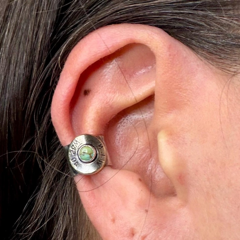 May include: A silver ear cuff with a circular design. The cuff has a small, round, light green stone in the centre. The metal is engraved with text around the edge. The cuff is worn on the ear.