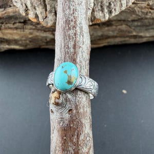 May include: A silver ring featuring an oval turquoise stone with brown inclusions. The ring band has a floral design. The ring is displayed on a wooden branch against a dark background.