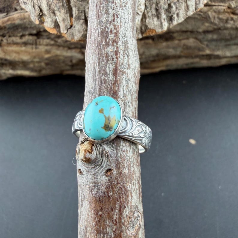 May include: A silver ring featuring an oval turquoise stone with brown inclusions. The ring band has a floral design. The ring is displayed on a wooden branch against a dark background.