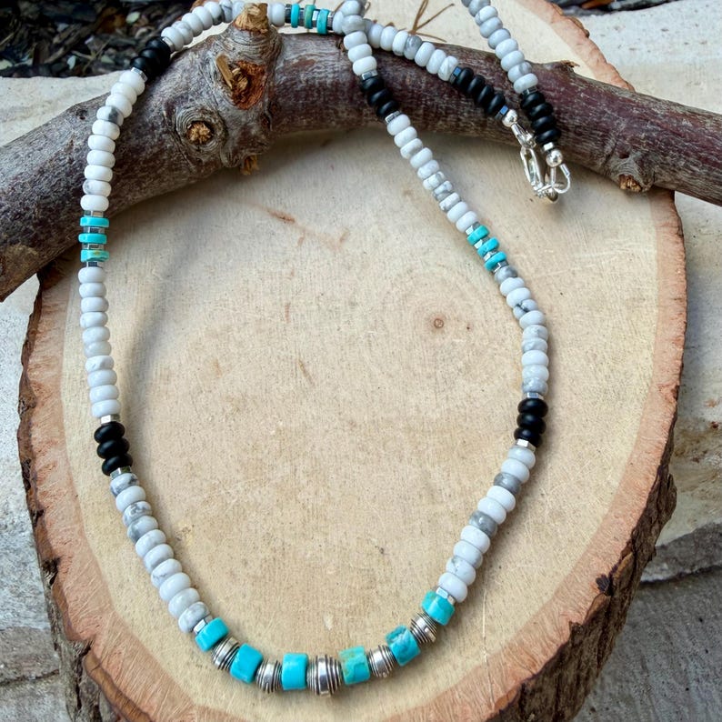 May include: A beaded necklace featuring white, gray, black, and turquoise beads. The necklace has a silver clasp and is displayed on a wooden surface. The beads are round and cylindrical, creating a unique pattern.