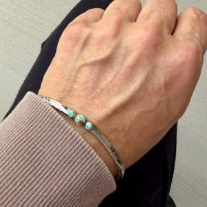 May include: A silver-toned cuff bracelet with a hammered texture. The bracelet is adorned with three turquoise-colored, round stones. The bracelet is shown on a person's wrist, with a neutral background.