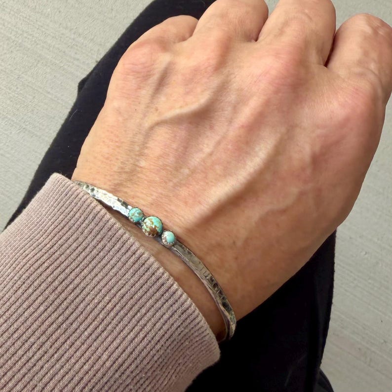 May include: A silver-toned cuff bracelet with a hammered texture. The bracelet is adorned with three turquoise-colored, round stones. The bracelet is shown on a person's wrist, with a neutral background.