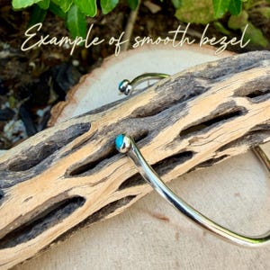 May include: A silver-toned bracelet with a smooth bezel, featuring a small, round, turquoise-colored stone at each end. The bracelet is displayed on a piece of weathered wood. The text "Example of smooth bezel" is written in cursive.