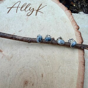May include: Four silver earrings with blue and white stones are displayed on a wooden branch. The earrings have different shapes, including oval and heart. The branch rests on a light-colored wooden surface. The name "Allyk" is written in cursive at the top.