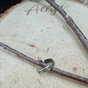 K&ouml;nnte beinhalten: Ein silberfarbener Ohrring aus Metall mit einem gepr&auml;gten Blattmuster auf dem &auml;u&szlig;eren Band. Das Schmuckst&uuml;ck ist auf einem d&uuml;nnen, braunen Holzzweig platziert, wobei der Text Ally K im Hintergrund auf einer hellen Holzoberfl&auml;che zu sehen ist.