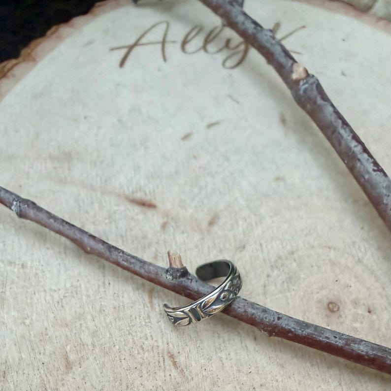 K&ouml;nnte beinhalten: Ein silberfarbener Ohrring aus Metall mit einem gepr&auml;gten Blattmuster auf dem &auml;u&szlig;eren Band. Das Schmuckst&uuml;ck ist auf einem d&uuml;nnen, braunen Holzzweig platziert, wobei der Text Ally K im Hintergrund auf einer hellen Holzoberfl&auml;che zu sehen ist.
