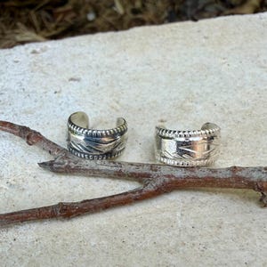 May include: Two silver-colored ear cuffs featuring a wide band design with a textured, repeating pattern along the center and a beaded border detail on the top and bottom edges. The metal has a polished finish with darkened recessed areas.