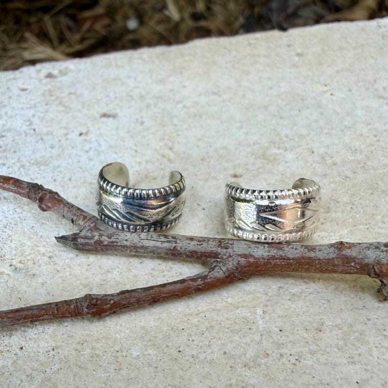 May include: Two silver-colored ear cuffs featuring a wide band design with a textured, repeating pattern along the center and a beaded border detail on the top and bottom edges. The metal has a polished finish with darkened recessed areas.