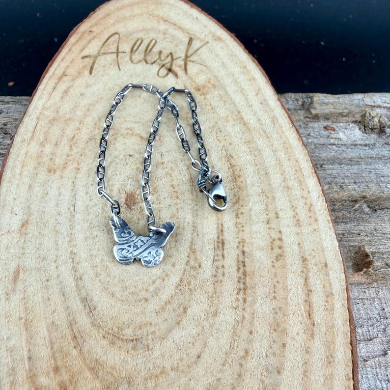 Op de afbeelding: Een zilveren vlinderhanger ketting met een ketting. De vlinderhanger heeft ingewikkelde ge&euml;tste details. De ketting wordt weergegeven op een houten oppervlak met de handgeschreven tekst "Ally K".