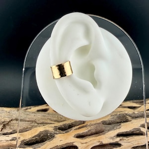 Puede incluir: Un brazalete de oreja dorado con textura martillada se muestra en un modelo de oreja blanco. El brazalete tiene un dise&ntilde;o de banda simple, mostrando un estilo minimalista. El fondo es oscuro, resaltando la joya.