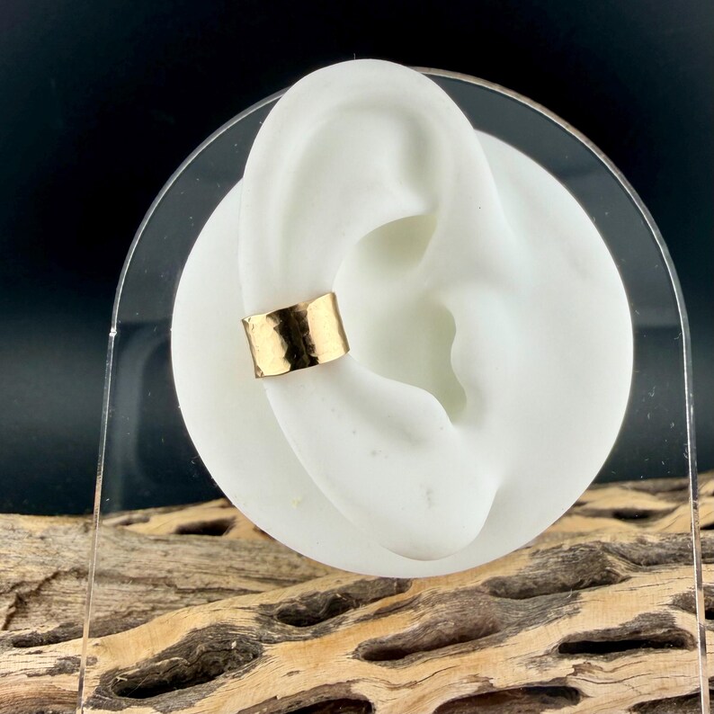Puede incluir: Un brazalete de oreja dorado con textura martillada se muestra en un modelo de oreja blanco. El brazalete tiene un dise&ntilde;o de banda simple, mostrando un estilo minimalista. El fondo es oscuro, resaltando la joya.
