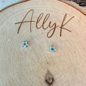 May include: A pair of star-shaped stud earrings with a silver-colored outline and a teal-colored center. The earrings are displayed on a wooden surface with the handwritten text "Allyk".