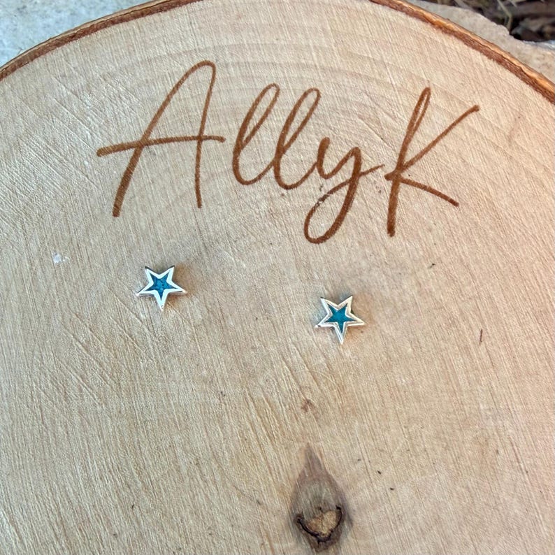 May include: A pair of star-shaped stud earrings with a silver-colored outline and a teal-colored center. The earrings are displayed on a wooden surface with the handwritten text "Allyk".