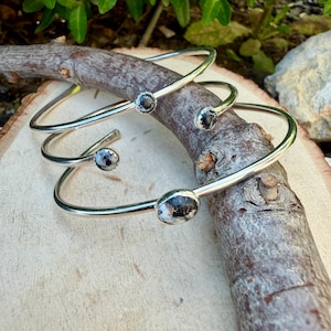 May include: Three silver-toned bangles with oval and round black and white speckled stones. The bangles are displayed on a wooden branch, showcasing their simple yet elegant design. The jewelry is suitable for everyday wear.