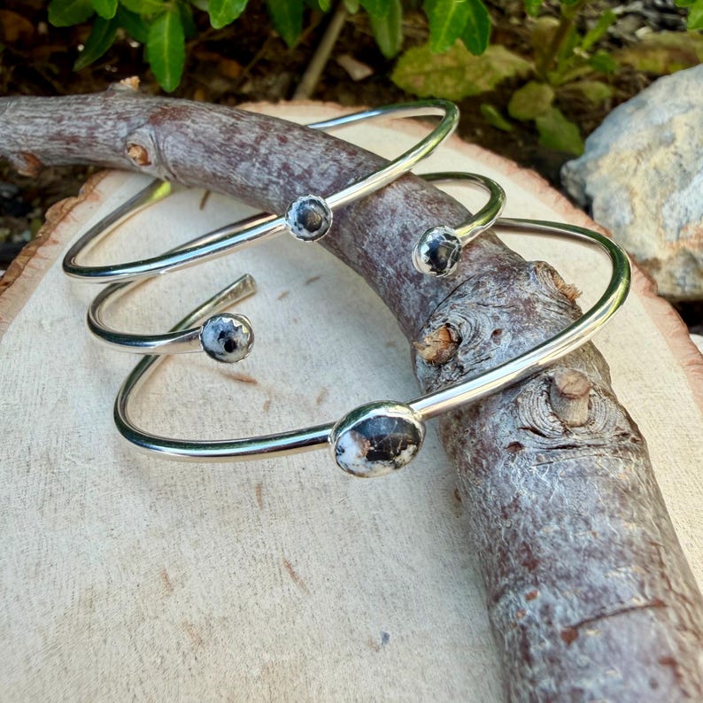 May include: Three silver-toned bangles with oval and round black and white speckled stones. The bangles are displayed on a wooden branch, showcasing their simple yet elegant design. The jewelry is suitable for everyday wear.