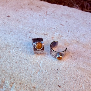 K&ouml;nnte beinhalten: Zwei Ringe aus silberfarbenem Metall mit einem breiten, strukturierten Band und eingravierten Mustern. Jeder Ring ist mit einem einzelnen, runden, polierten Tigerauge-Edelstein in warmem Goldbraun besetzt. Die Ringe sind offen.