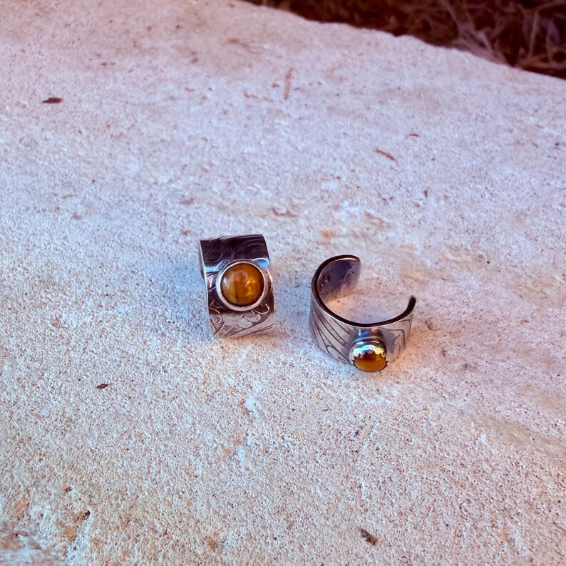 K&ouml;nnte beinhalten: Zwei Ringe aus silberfarbenem Metall mit einem breiten, strukturierten Band und eingravierten Mustern. Jeder Ring ist mit einem einzelnen, runden, polierten Tigerauge-Edelstein in warmem Goldbraun besetzt. Die Ringe sind offen.