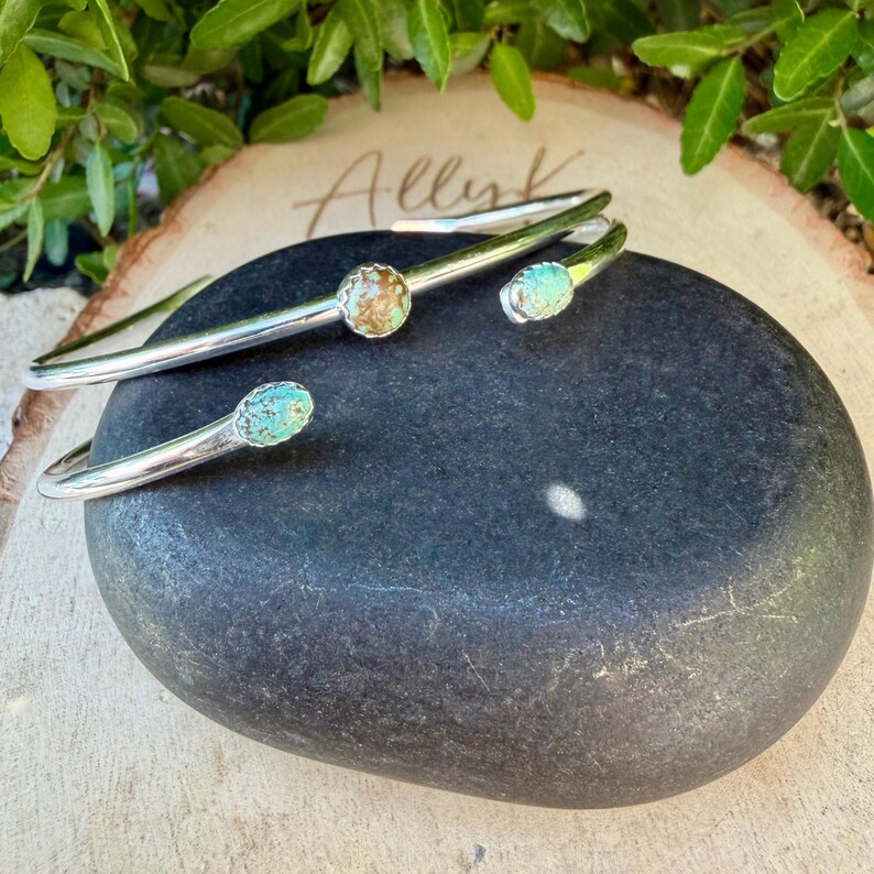 May include: Three silver-toned cuff bracelets with turquoise-colored stone accents. The bracelets are displayed on a dark gray stone. The background includes a wooden surface with the handwritten name "Allyk" and green foliage.
